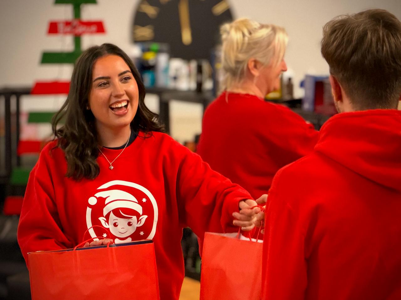 NBCC volunteers in red hoodies sharing a laugh as they hand over Red Bags Full of Joy at the Elves Workshop